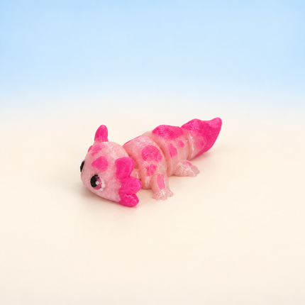 Micro Axolotl 3D-printed miniature featuring a pink body, feathery external gills, and a long tail, on a light surface with a pale blue background.
