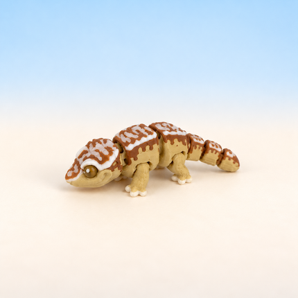 Micro Crested Gecko 3D-printed miniature featuring a patterned brown and cream body and a long tail, on a light surface with a pale blue background.