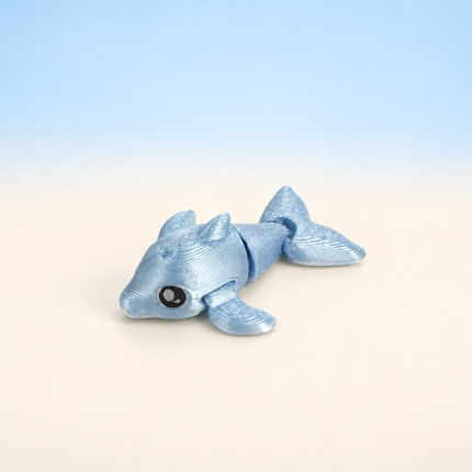 Micro Dolphin 3D-printed miniature featuring a blue-gray body, curved fins, and a smiling face, on a light surface with a pale blue background.