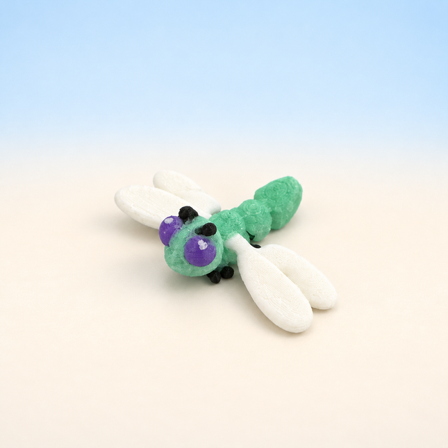 Micro Dragonfly 3D-printed miniature featuring a green body, large eyes, and pale outstretched wings, on a light surface with a pale blue background.