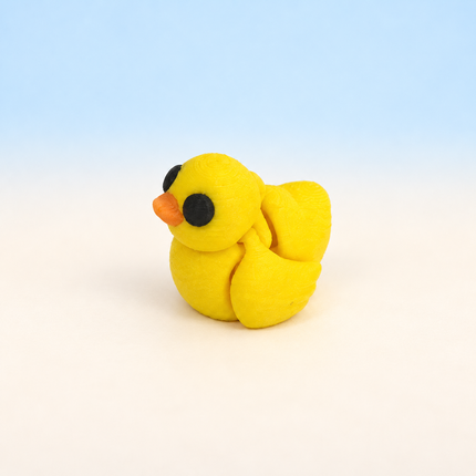 Micro Duck 3D-printed miniature featuring a yellow body, orange beak, and rounded wings, on a light surface with a pale blue background.