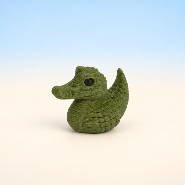 Micro Gator Duck 3D-printed miniature featuring a green duck-shaped body with an alligator-inspired snout and textured back, on a light surface with a pale blue background.
