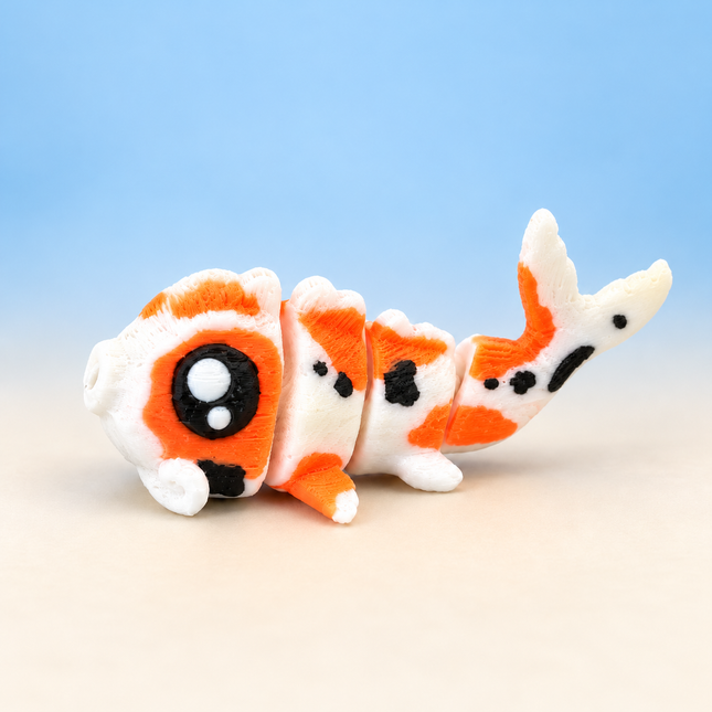Micro Koi 3D-printed miniature with an orange, white, and black patterned body with flowing fins, on a light surface against a pale blue background.