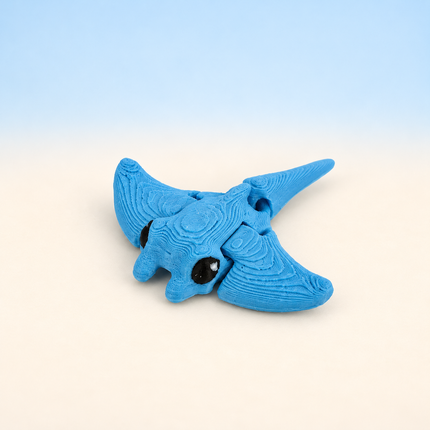 Micro Manta Ray 3D-printed miniature featuring a blue body, broad wing-like fins, and a long tail, on a light surface with a pale blue background.