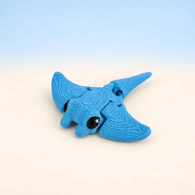Micro Manta Ray 3D-printed miniature featuring a blue body, broad wing-like fins, and a long tail, on a light surface with a pale blue background.