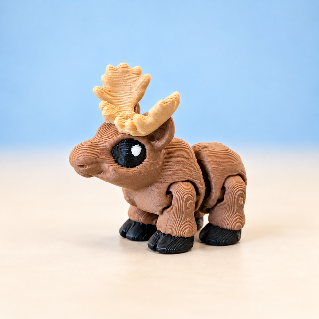 Micro Moose 3D-printed miniature with a brown body, broad tan antlers, and dark hooves, on a light surface against a pale blue background.